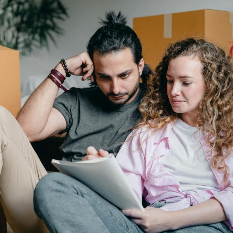 Pensive male and female in casual clothes sitting together among boxes and writing notes in notebook while leaning on bed